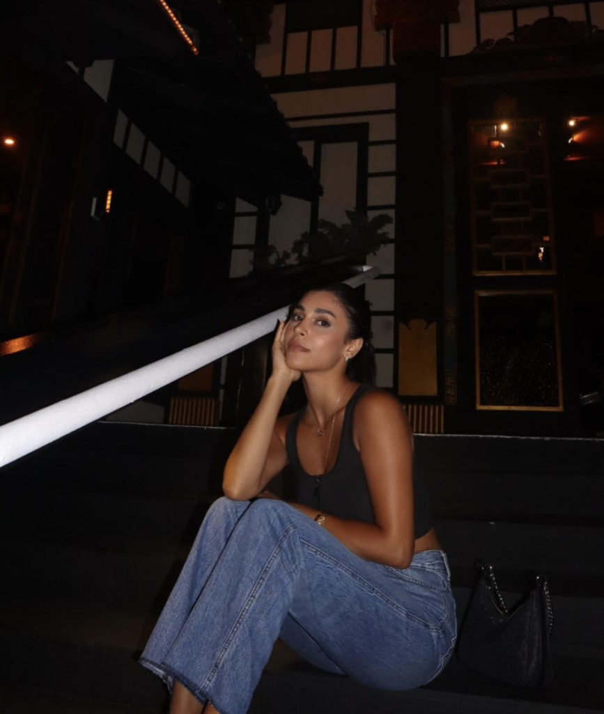 Woman sitting on steps at night in a tank top and jeans, and a black purse beside her.